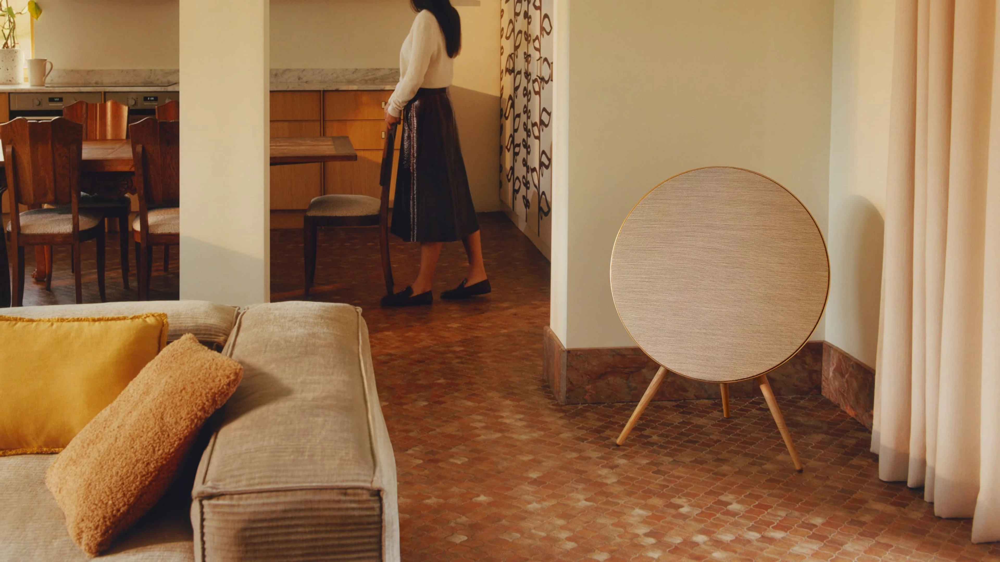 Beosound A9 in a living room corner next to a sofa, on the back there is a lady in the kitchen