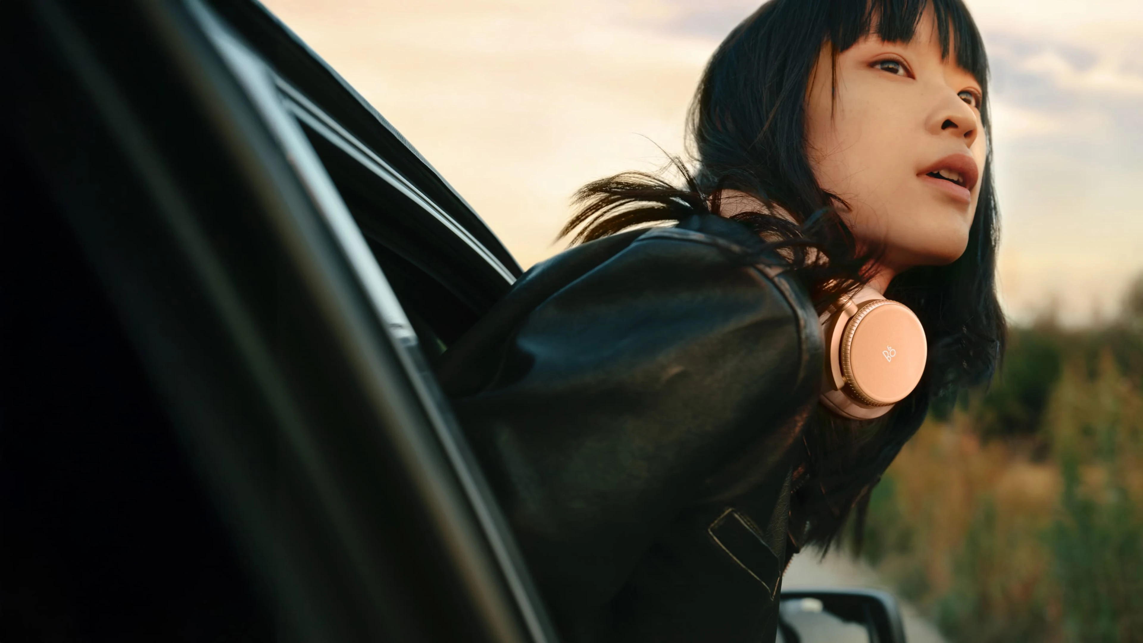 A woman putting her head out of the car window and she has Sunset Apricot color Beoplay H100 around her neck